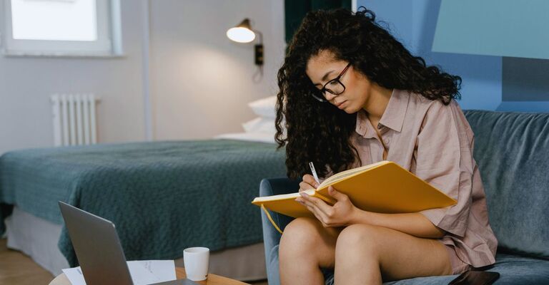 Ragazza riccia con gli occhiali seduta sul divano che pende appunti su un notebook color arancio