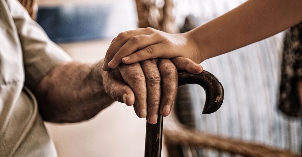 Mano di un'anziana persona appoggiata a un bastone, coperta con delicatezza dalla mano di un bambino, simbolo di connessione tra generazioni e fragilità dell'invecchiamento.