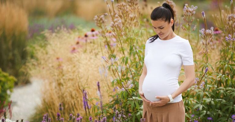 Antiacidi in gravidanza Donna incinta in piedi in un giardino fiorito, con le mani sul pancione e lo sguardo rivolto verso il basso.