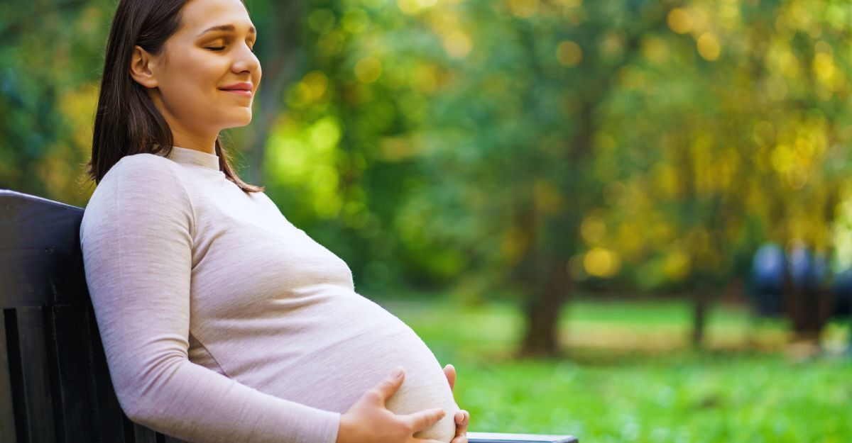 Donna incinta seduta su una panchina nel parco, con espressione serena e le mani sul pancione.