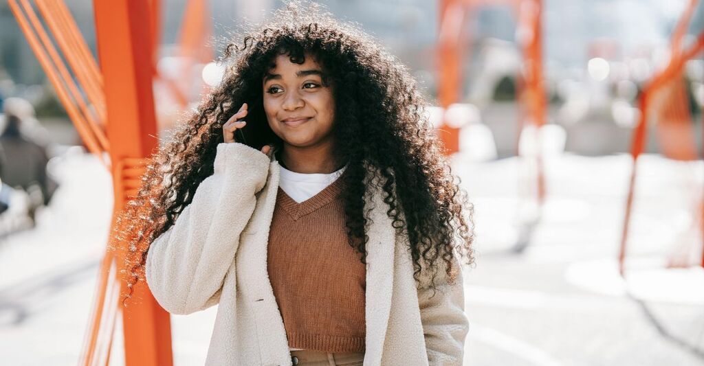 Ragazza riccia e sorridente mentre cammina per strada e parla al telefono