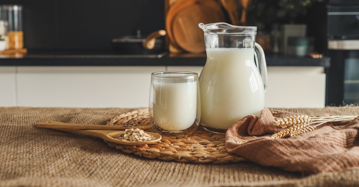 Una brocca e un bicchieri pieni di latte di avena sul tavolo in cucina, circondati da semi e spighe di avena su uno strofinaccio