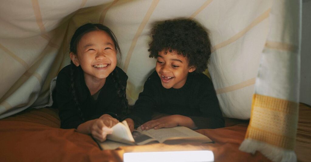 Due bambini in una tenda fatta di coperte che leggono e ridono insieme grazie alla luce di una torcia