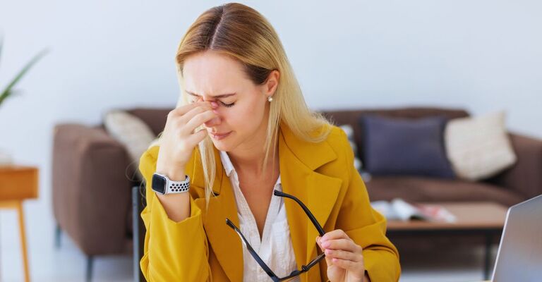 Donna seduta alla scrivania mentre usa il PC che si tiene il setto nasale con le dita a occhi chiusi mentre stringe in mano degli occhiali da vista neri