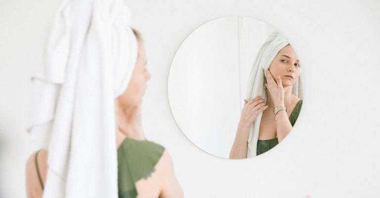 Ragazza che si guarda allo specchio tondo mentre si spalma una crema dopo la doccia, con un asciugamano in testa