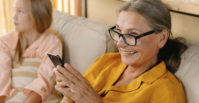 Signora, seduta sul divano accanto alla nipote, con il cellulare in mano che sorride a qualcuno