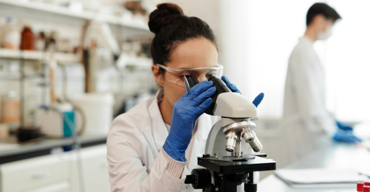 Ricercatrice in laboratorio che con occhiali specifici e guanti blu osserva al microscopio dei campioni 