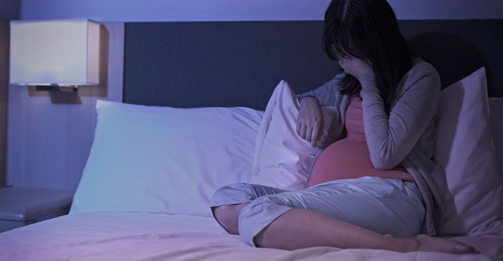 Donna incinta seduta sul letto di notte con una mano sul volto in segno di stanchezza o tristezza.