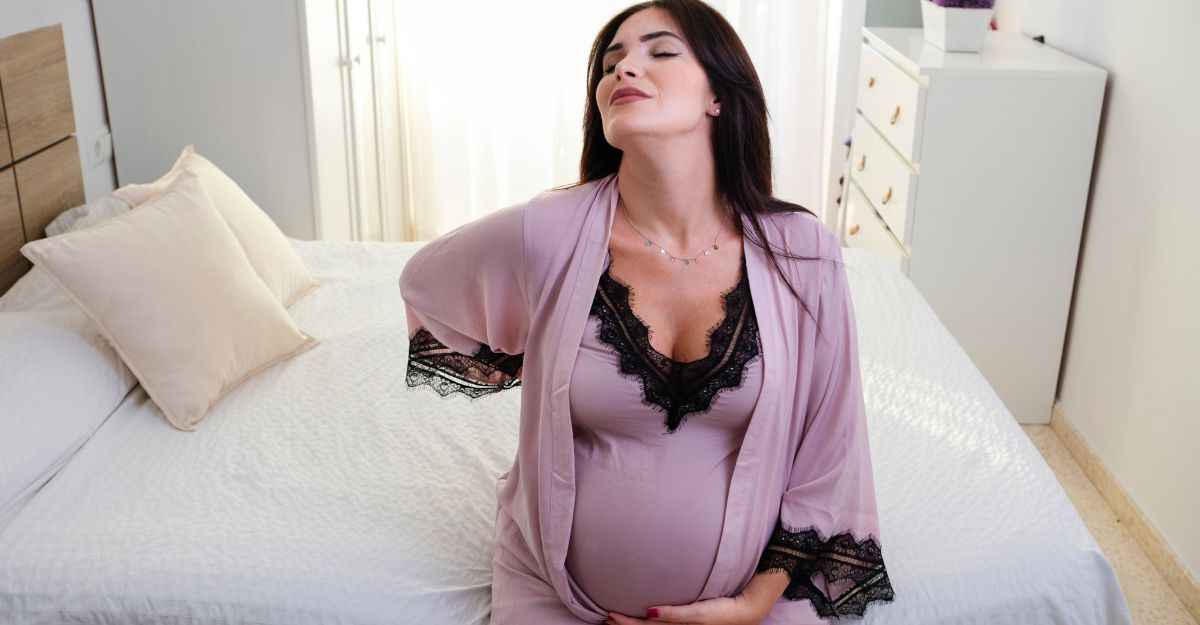 Donna incinta seduta sul letto con espressione di dolore e una mano sulla schiena, in camera da letto illuminata di giorno.