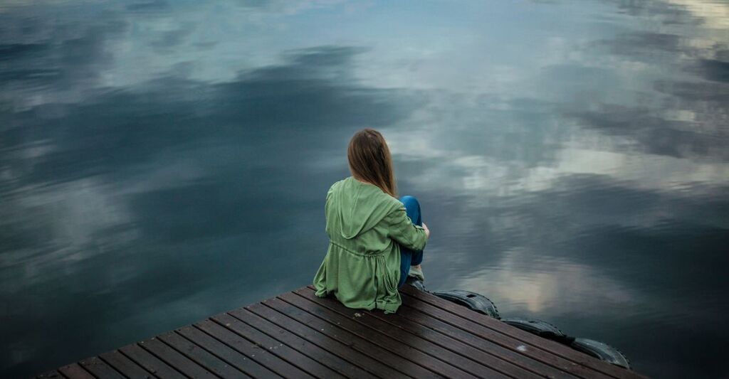 Una persona seduta su un molo di legno, di spalle, con indosso una giacca verde, guarda il riflesso del cielo sull’acqua calma, in un momento di introspezione e solitudine.