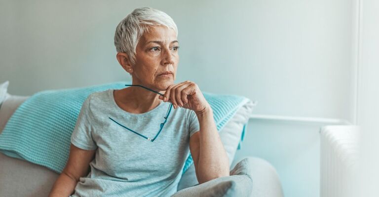 Donna di mezza età con i capelli bianchi e corti seduta sul divano con gli occhiali in mano che guarda fuori dalla finestra