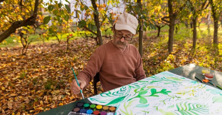 Uomo anziano che dipinge con acquerelli in un parco autunnale, esempio di creatività e tempo libero riscoperto in pensione.