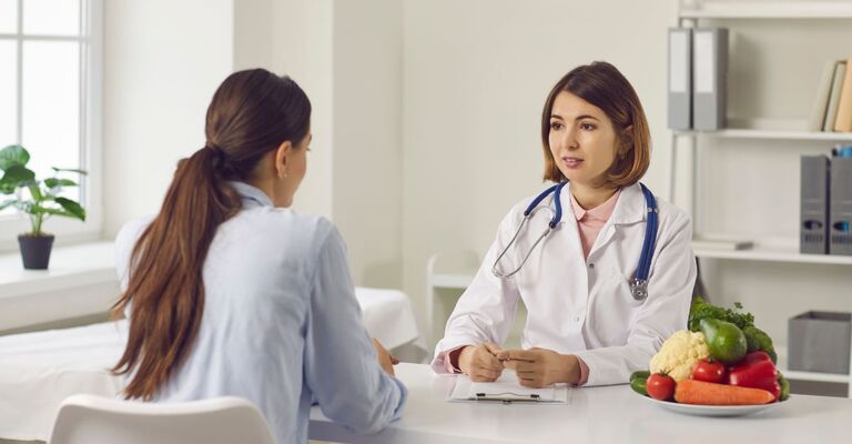 Medico nutrizionista parla con una paziente in ambulatorio, con un piatto di verdure fresche sul tavolo per discutere di alimentazione e salute.