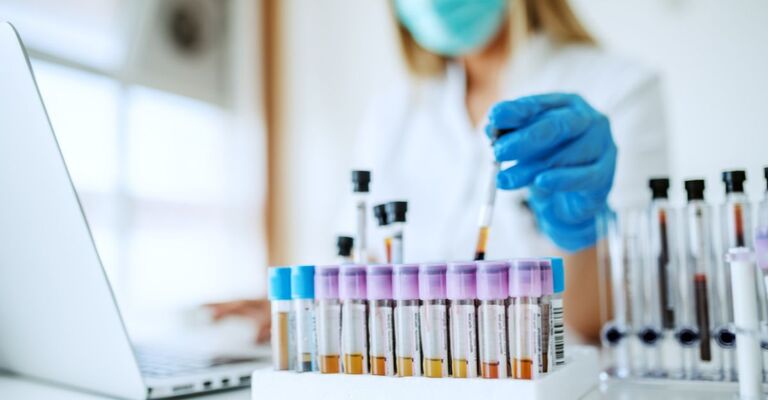 Primo piano di fialette di campioni di laboratorio mentre una scienziata le maneggia con i guanti e la mascherina davanti al PC