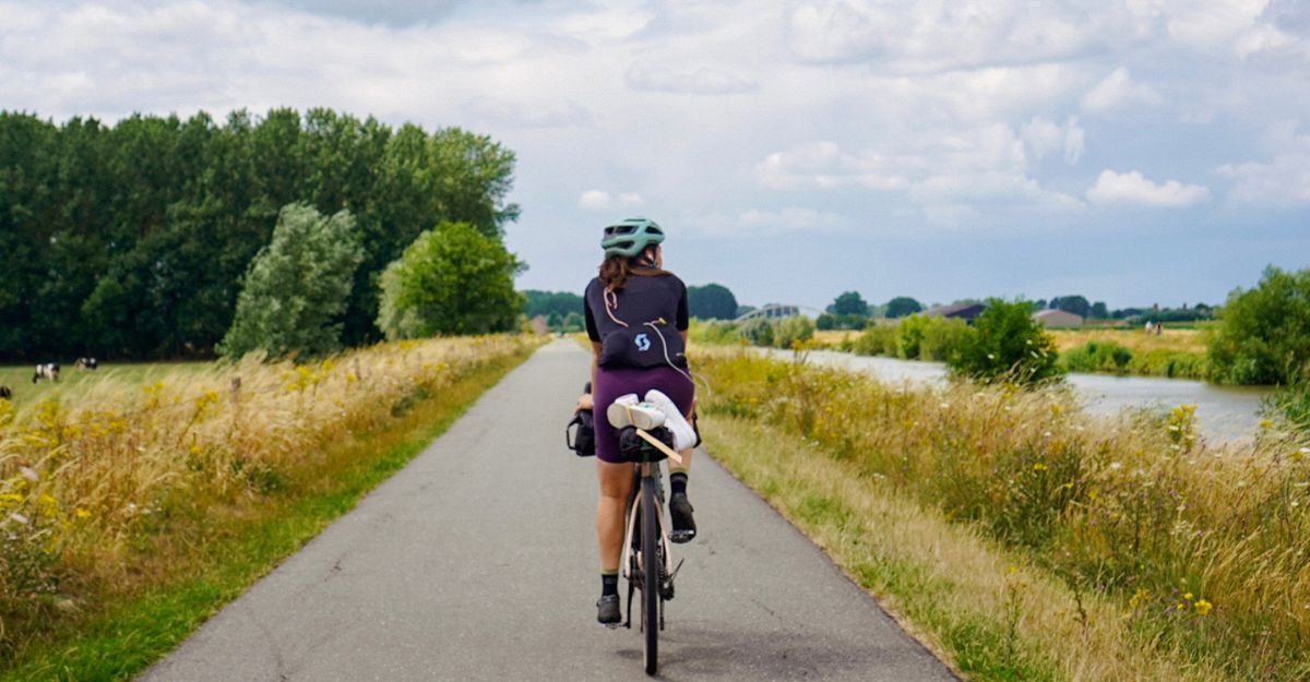 ragazza in bicicletta, attraversa una strada in mezzo ai campi e ad un corso d'acqua