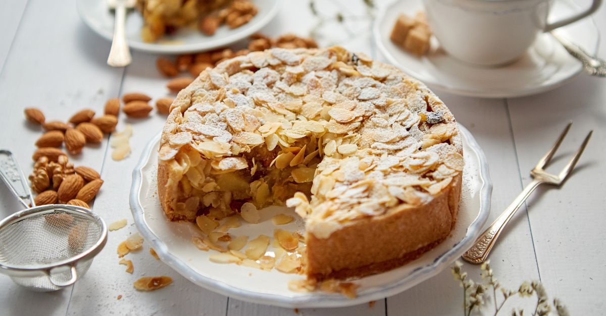 Torta di mandorle ricoperta da scaglie di mandorle con accanto una forchettina e una tazza di cappuccino