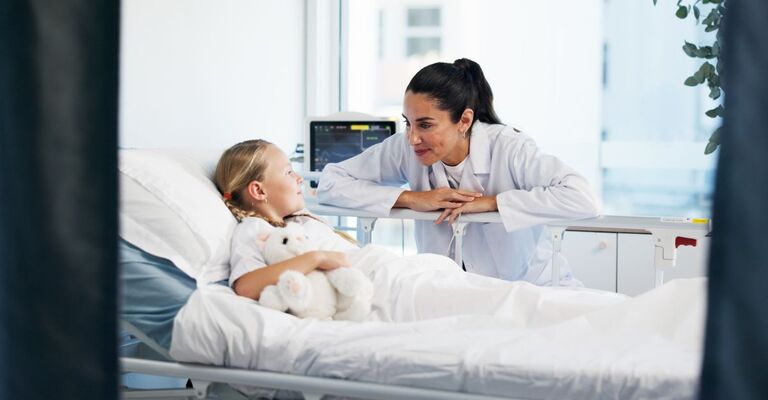 Pediatra in reparto appoggiata al letto che sorride a una bambina sdraiata nella stanza d'ospedale con un orsacchiotto