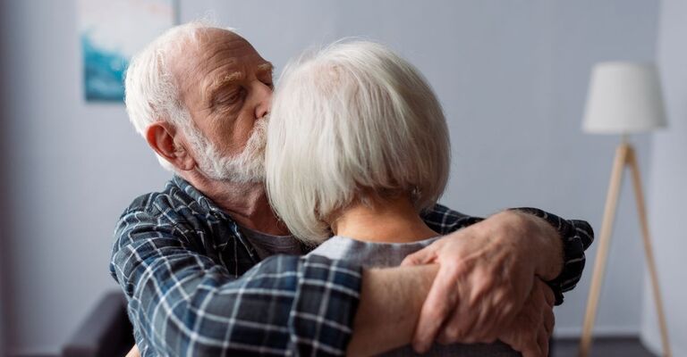 Un uomo anziano abbraccia e bacia una donna anziana sulla testa, con le braccia avvolte intorno a lei in modo protettivo.