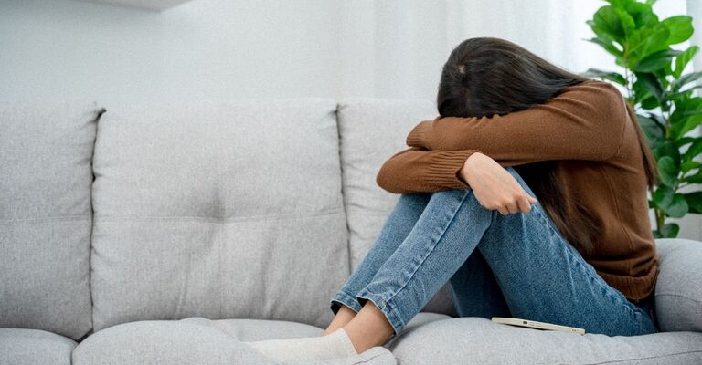 Una donna seduta su un divano, con le ginocchia al petto e il viso nascosto tra le braccia, in una posizione che suggerisce tristezza o disperazione.