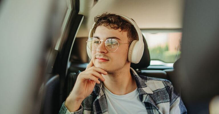Ragazzo in auto seduto dietro che ascolta la musica con delle cuffie grandi e bianche mentre sorride e guarda fuori dal finestrino