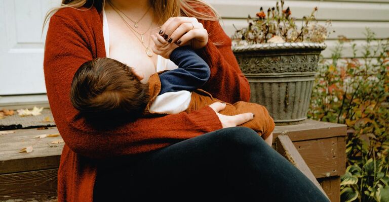 Primo piano di donna con cardigan rosso che allatta il figlio seduta sulle scale del porticato di casa