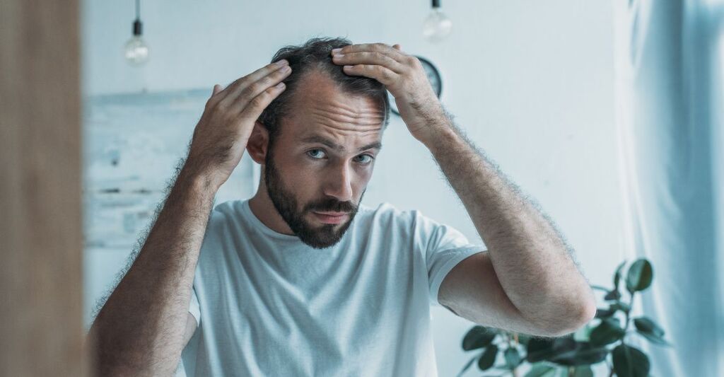 Uomo con barba che osserva la propria testa toccando i capelli diradati davanti allo specchio, preoccupato per la caduta.