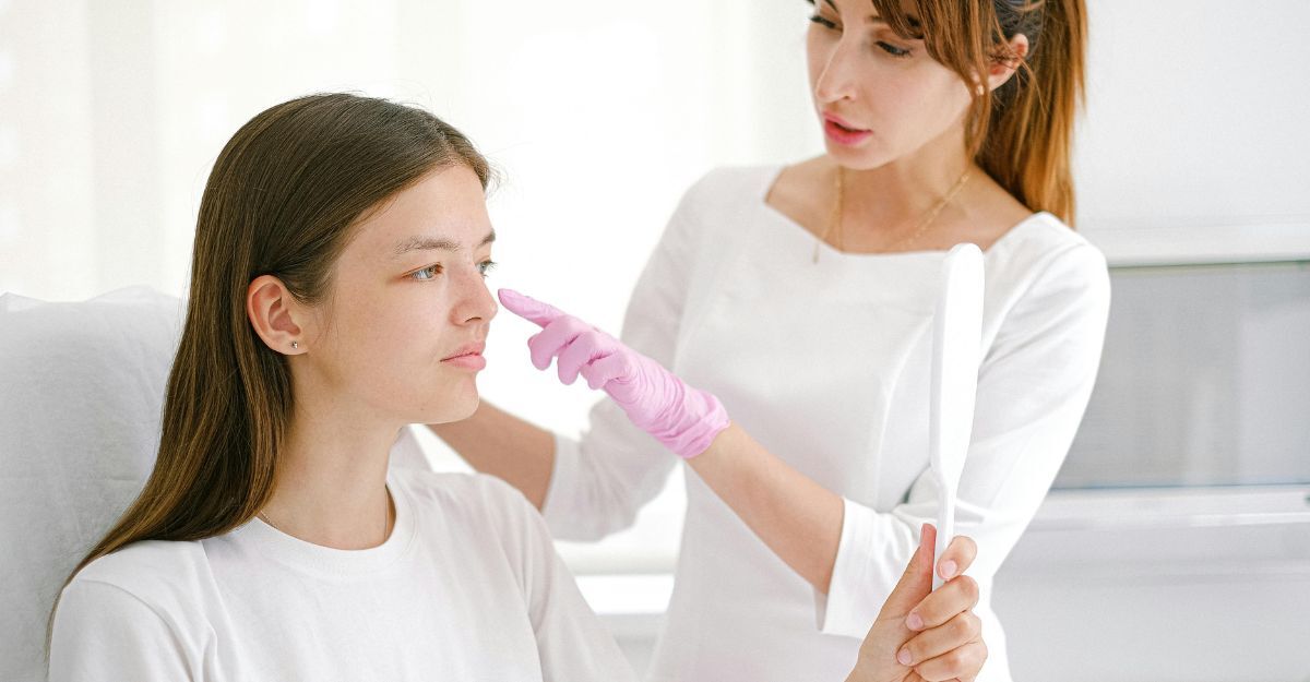 Una dermatologa in camice bianco e guanti rosa tocca il naso di una giovane paziente, mentre la paziente si guarda allo specchio.