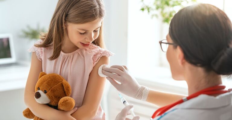 Una bambina con in braccio un orsacchiotto guarda un'infermiera che le sta per fare una puntura al braccio.