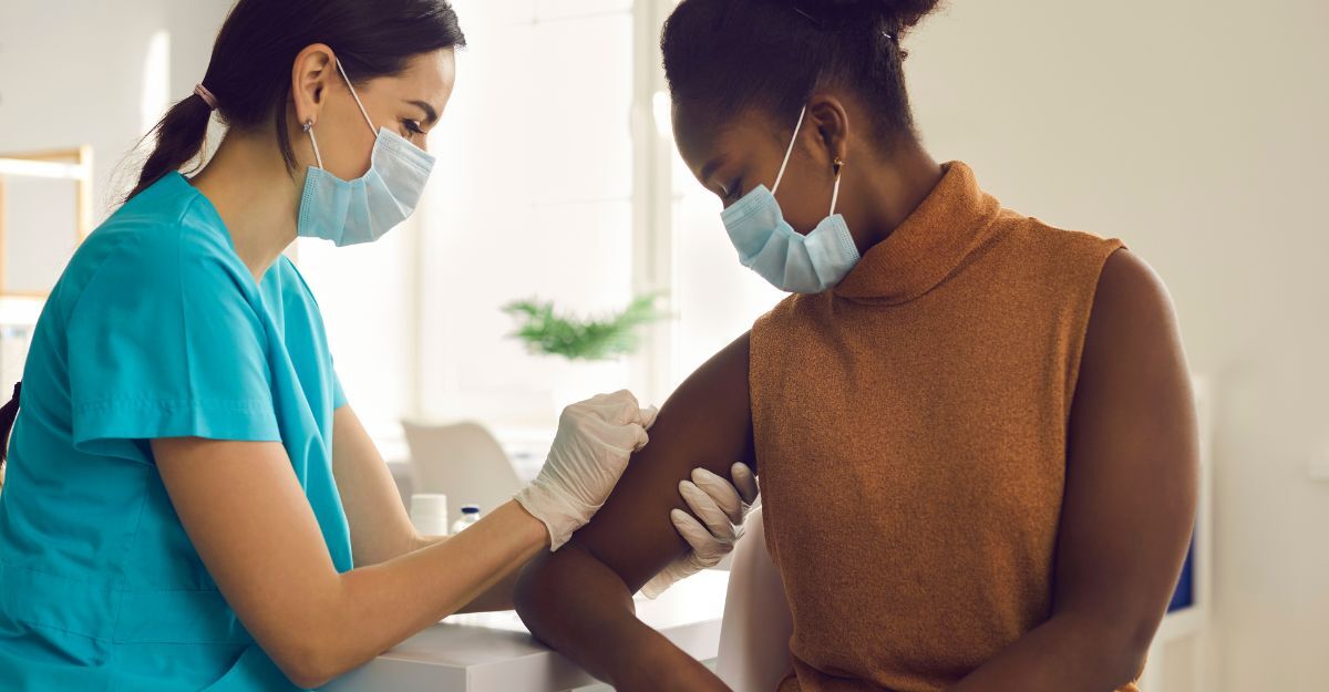 Un'infermiera con maschera e guanti, in camice azzurro, sta per fare una puntura sul braccio di un'altra persona. Entrambi indossano la mascherina.