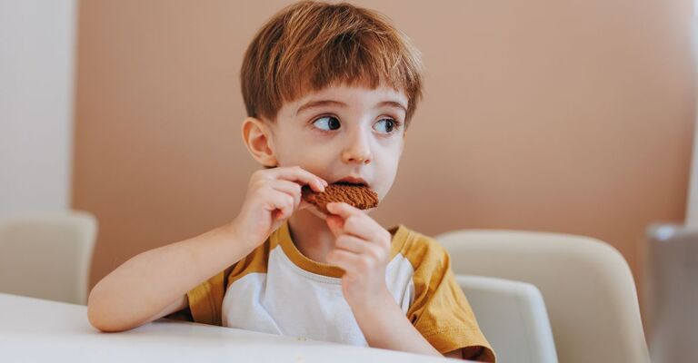 Un bambino seduto a tavola che guarda di lato mentre mangia un biscotto.