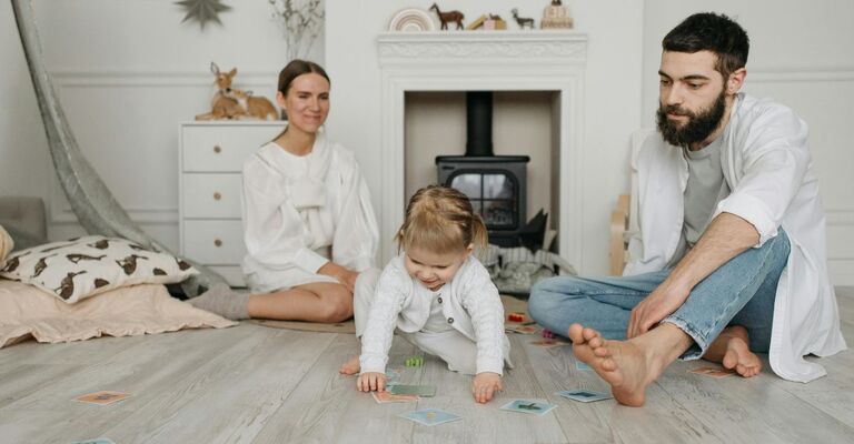 Neonata che gattona sul pavimento vicino ai due genitori e a delle carte per imparare nella sua cameretta