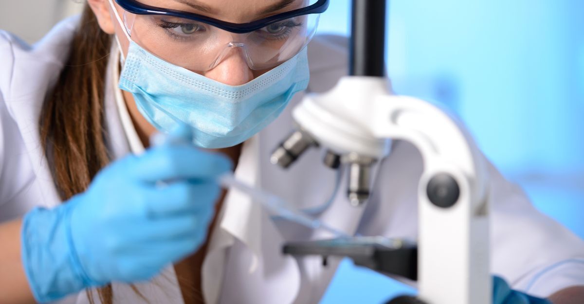 Primo piano di una scienziata con mascherina e occhiali di protezione, che lavora con un microscopio in laboratorio.