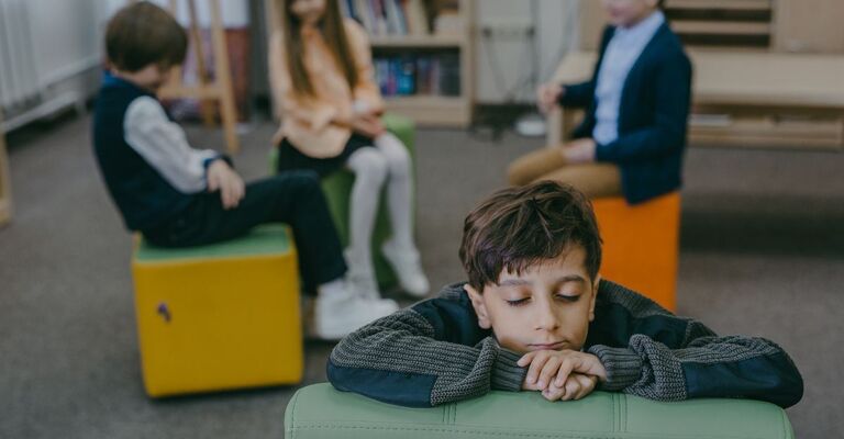 Un bambino si appoggia tristemente a un tavolino in una biblioteca o in un'aula, isolato dagli altri bambini che si vedono sullo sfondo.