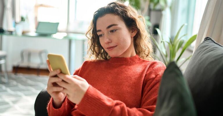 Donna con maglione rosso che chatta sullo smartphone con aria dubbiosa