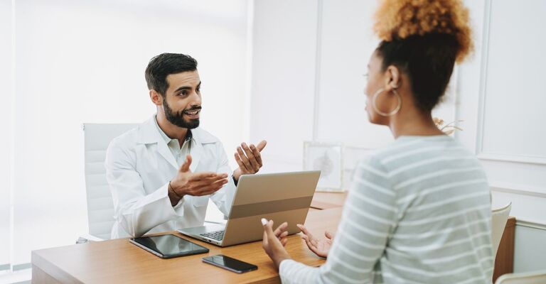 Un medico con uno stetoscopio al collo è seduto su un divano e parla con una paziente, anche lei seduta di fronte a lui.