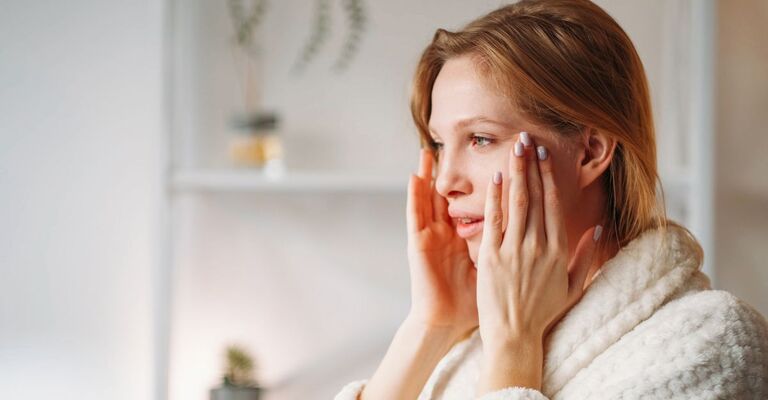 Giovane donna bionda con occhi azzurri in accappatoio di spugna bianco che tasta il suo volto con le mani