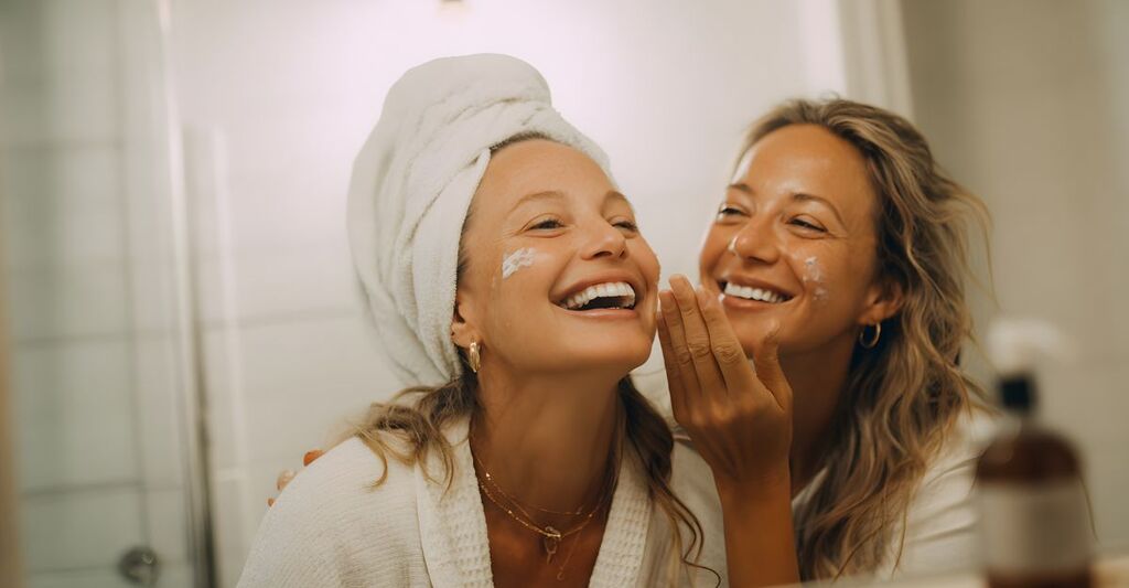 Due donne adulte e sorridenti sono in un bagno, con asciugamani in testa e del prodotto idratante applicato sul viso.