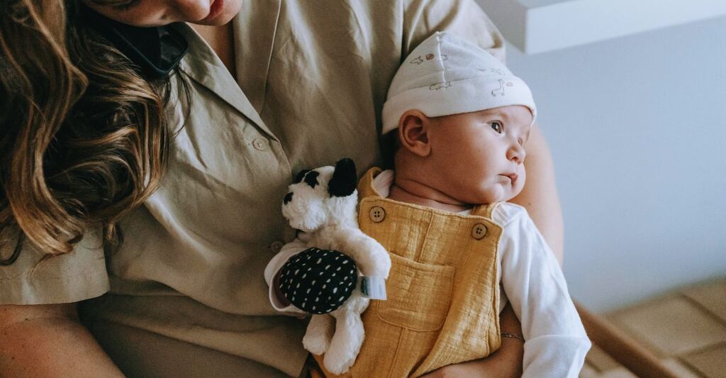 Primo piano di un neonato appoggiato sulla mamma con un cane di peluche sopra la salopette color senape