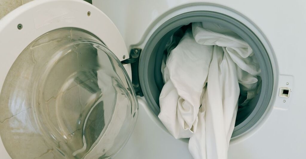 Primo piano di un oblò di una lavatrice aperto con delle lenzuola bianca che sbucano