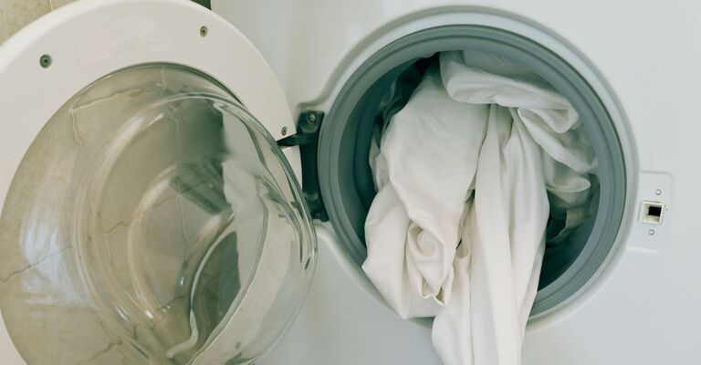 Come lavare le lenzuola in lavatrice: guida completa e consigli pratici Primo piano di un oblò di una lavatrice aperto con delle lenzuola bianca che sbucano
