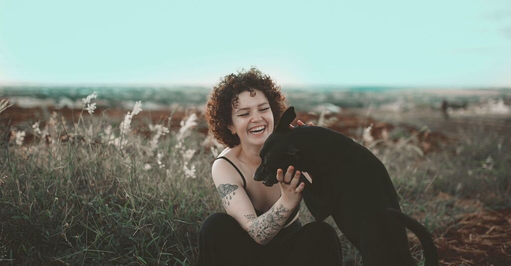 Donna con tatuaggi e canotta che sta coccolando il suo cane dal manto nero al parco