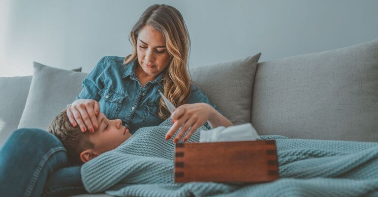Mamma e figlio sul divano. Il figlio ha la febbre alta, la coperta e una scatola di fazzoletti addosso, e la mamma gli mette una mano in fronte per rassicurarlo.