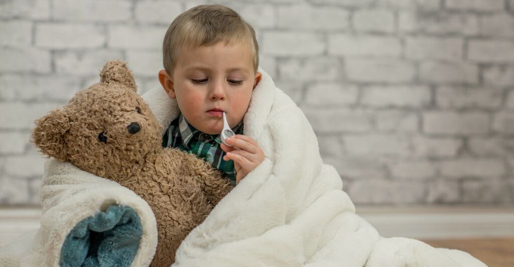 Un bambino avvolto da una coperta con un pupazzo e il termometro in bocca