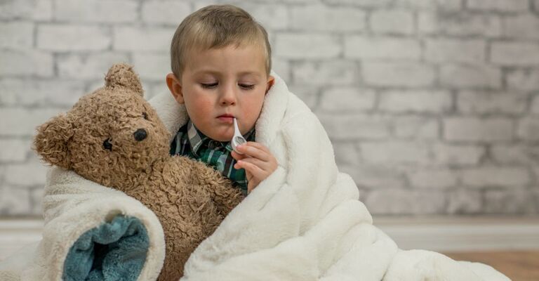 Long Covid bambini Un bambino avvolto da una coperta con un pupazzo e il termometro in bocca