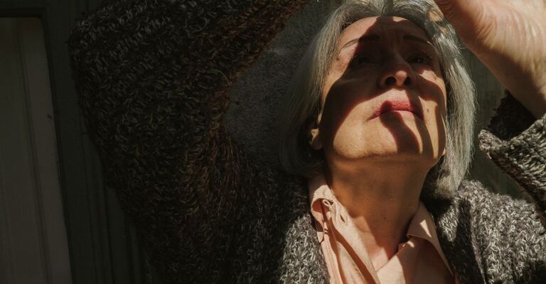 Un'anziana signora dai capelli grigi solleva il braccio, in piedi in penombra, il viso in parte coperto da ombre nette.