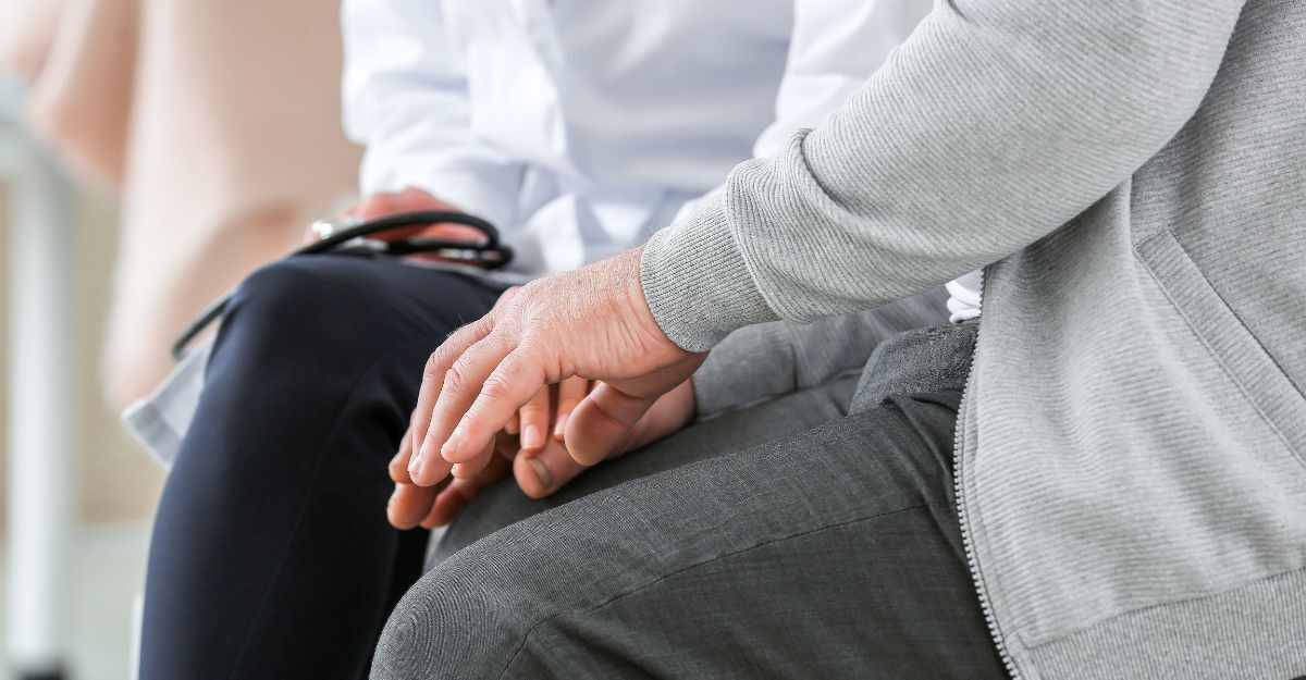 Primo piano sul tronco e le mani intrecciate di un paziente anziano seduto accanto a una persona che indossa un camice da medico.