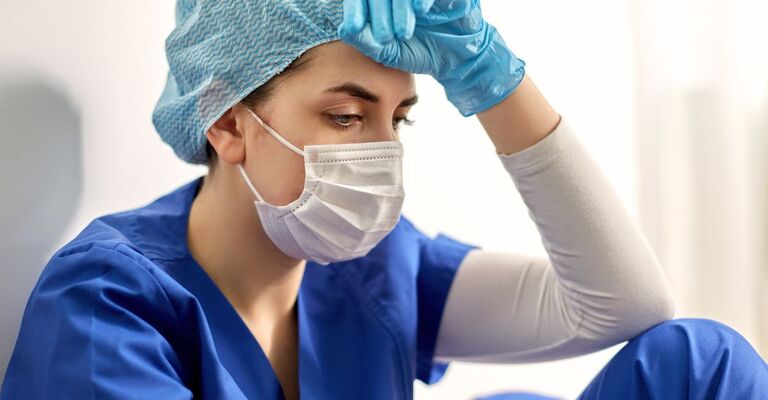 Primo piano di operatrice sanitaria con camice, cuffia chirurgica e mascherina, la mano guantata appoggiata sulla fronte.