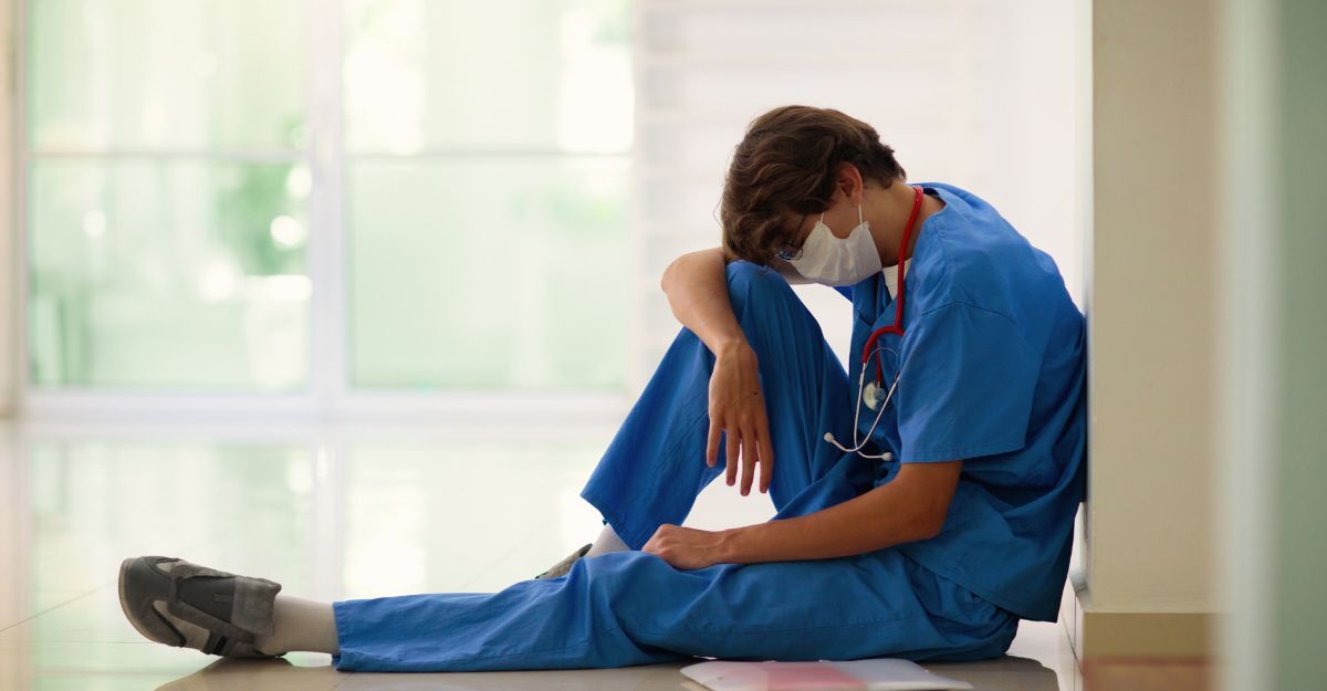 Operatore sanitario in camice blu e mascherina seduto a terra, testa tra le mani.