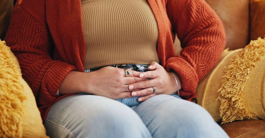 Mani, dolore e persona con crampi allo stomaco, stitichezza e sindrome dell'intestino irritabile sul divano. Casa, malattia e donna con malattia infiammatoria pelvica, mestruazioni e tocco della pancia