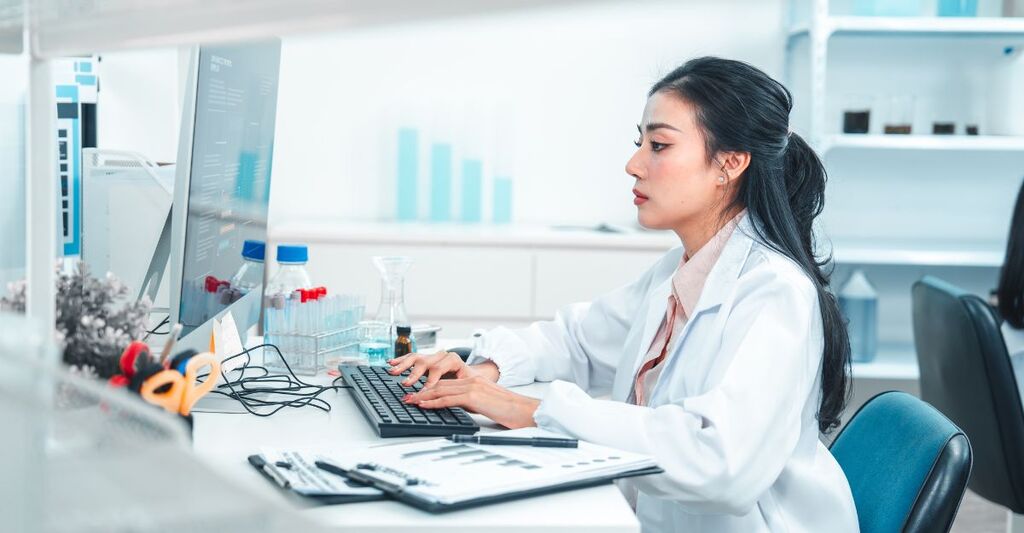 Una scienziata in camice da laboratorio digita su una tastiera mentre analizza i dati di una ricerca al computer. Sono visibili attrezzature di laboratorio e campioni chimici, a dimostrazione di un ambiente di lavoro scientifico e concentrato